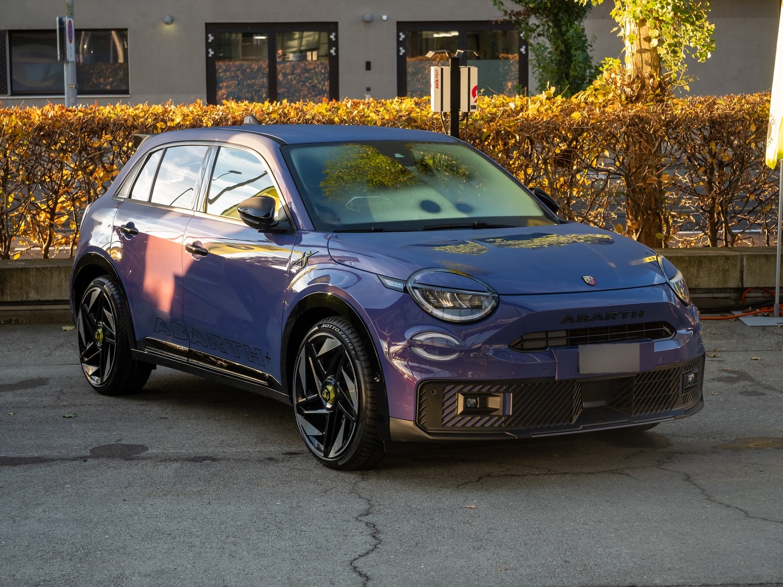 Abarth 600e viola, vista anteriore di tre quarti davanti a una siepe autunnale